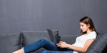Young student girl in white t-shirt and blue jeans works on her laptop computer laying on grey coach sofa in front of grey wall