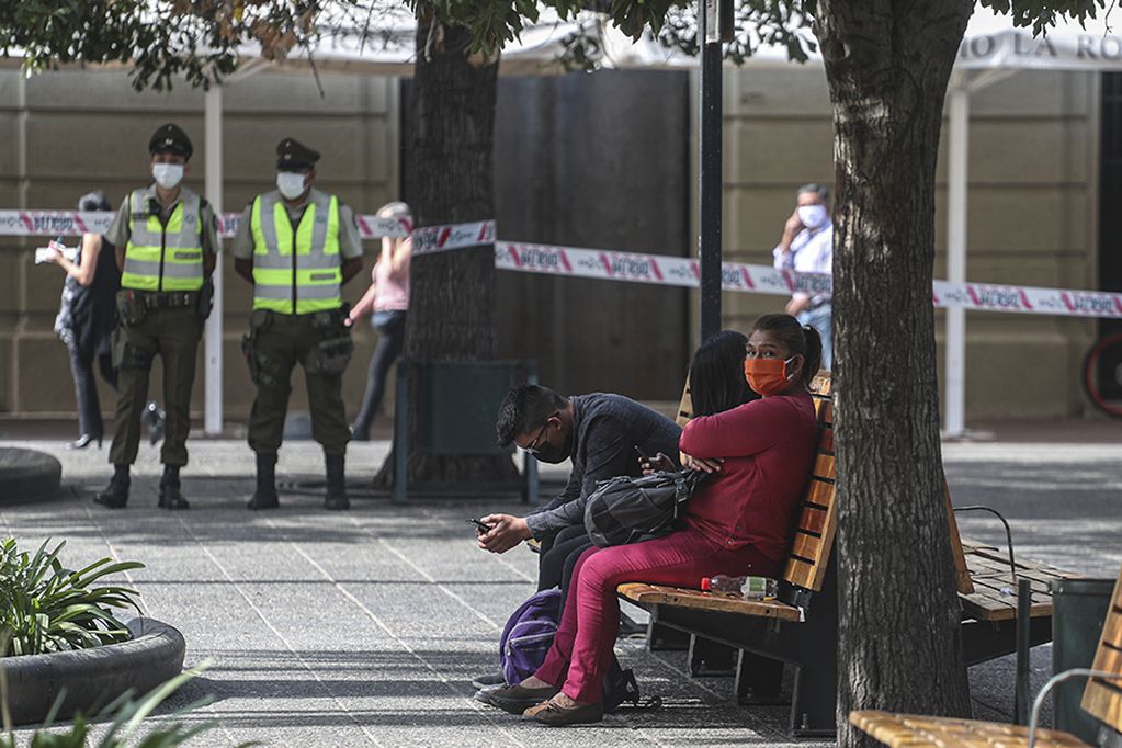 Police stand guard over a woman, right, who tested positive for the new coronavirus, sits on a park bench accompanied by her son and his girlfriend, in Santiago, Chile, Wednesday, April 15, 2020. The 49-year-old woman, a Peruvian national and resident in Chile, broke her mandatory quarantine on Wednesday and visited the Plaza de Armas to "stretch her legs" she said. (AP Photo/Esteban Felix)