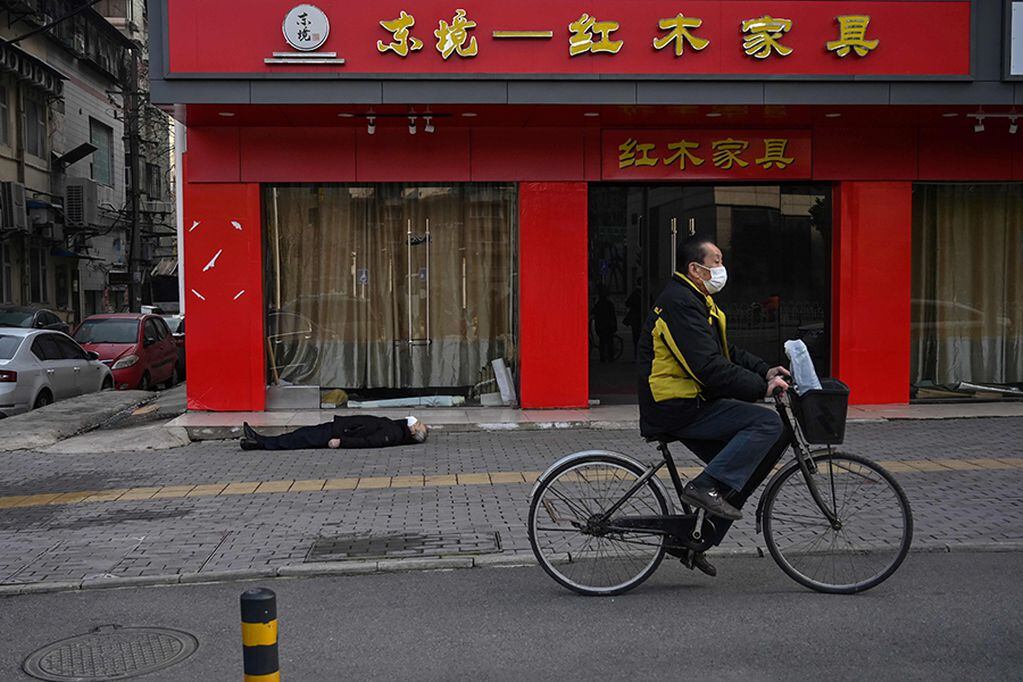 This photo taken on January 30, 2020 shows a man wearing a facemask cycling past an elderly man collapsed and died on the pavement along a street near a hospital in Wuhan. - AFP journalists saw the body on January 30, not long before an emergency vehicle arrived carrying police and medical staff in full-body protective suits. The World Health Organization declared a global emergency over the new coronavirus, as China reported on January 31 the death toll had climbed to 213 with nearly 10,000 infections. (Photo by Hector RETAMAL / AFP) / TO GO WITH China-health-virus-death,SCENE by Leo RAMIREZ and Sebastien RICCI