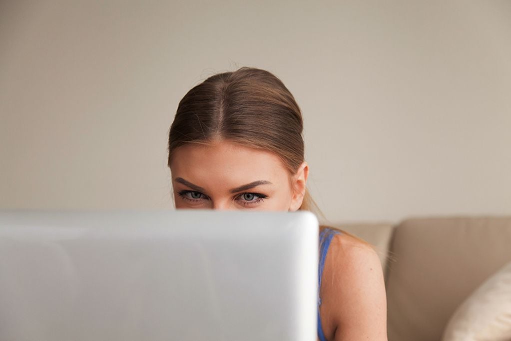 Curious teen using computer, young funny girl peeking over top of laptop, looking at screen, playing interesting online game, having fun while chatting with friend, secretly watching video at home
